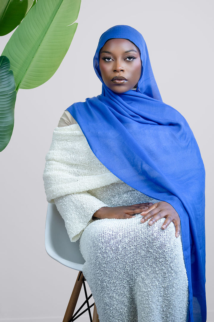Woman wearing a blue hijab and white outfit sitting on a chair with a plant in the background