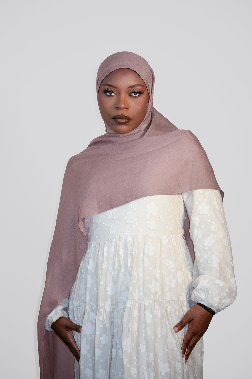 Woman wearing a light pink hijab and white dress against a plain background